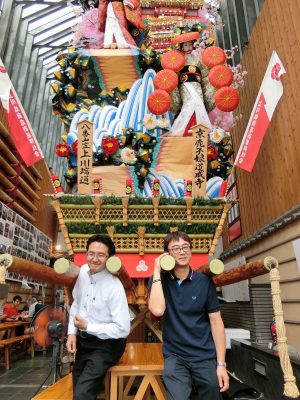 商店街に飾られた山笠を担いでみせる川端中央商店街「みどりや仏壇店」代表の吉川和毅さん(左)と上川端商店街「子供服jay's」代表の渡辺淳一郎さん。子どもの頃から別々の山笠を担いで競ってきたが、商店街の運営では力を合わせる
