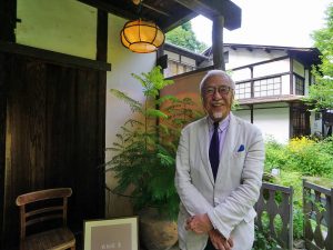 ▲娘婿の視点から次郎・正子夫妻の暮らしぶりを描いた著書も多数ある牧山圭男さん。NHKドラマスペシャル『白洲次郎』の中では実はエキストラとしても登場したそう。「映ったのはほんの5秒ぐらいだけどね」と笑う牧山さんは次郎氏の姿を連想させるチャーミングな紳士だ。牧山さんの右側背後に見えているのが旧白洲邸のダイニングだった建物(現在は『レストラン&カフェ武相荘』として活用中)