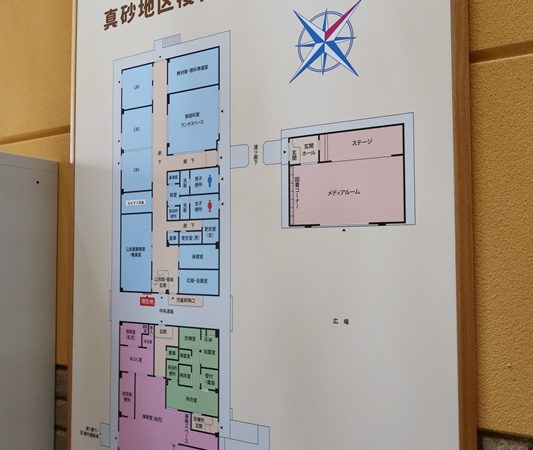 益田市立地域活性化交流館の玄関に掲げられた施設案内図。水色部分が小学校と公民館、ピンクの部分が保育園、緑が診療所、うすいピンクの部分がある意味、共有部的な空間