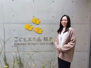 蝶を施したロゴマークの横に立つのは施設の案内をしていただいた「こども本の森 熊本」の主任主事・坂口沙采さん