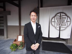 CSA不動産の代表取締役社長 小島 孝仁社長。後ろは、蔵を改造したイタリアン＆フレンチのお店「KURAYA KATO」