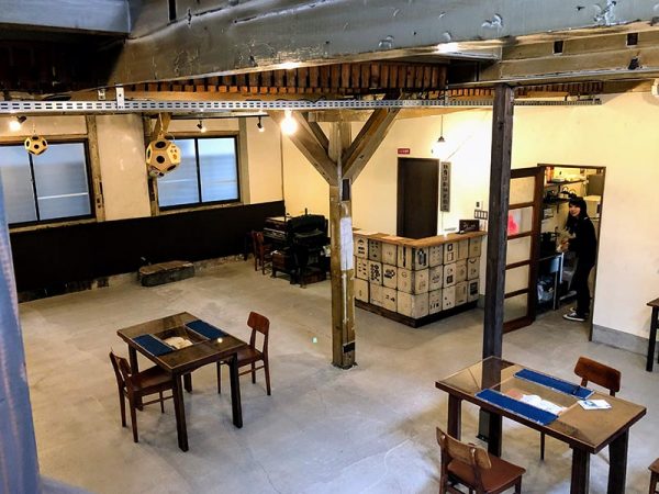 階段から玄関広間を俯瞰する。椅子はもともとここにあったもの。テーブルは工場の廃材を再利用してつくった。写真奥、開いているドアから覗いているのが知子さん。ドア横のコーナーが「番台」で、柄のついたタイルのようなものは、印刷に使っていた石版だ。番台の左に置いてあるのは、昔ながらの石版印刷機