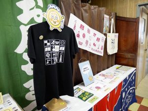 資料館では、Tシャツやトートバッグ、タオル、缶バッジなど、公認キャラクター「おけお」グッズが展示販売されている