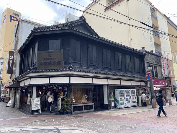 築町商店街の角地に建つ小野原本店。入母屋屋根の頂上の鬼瓦には「小」の文字が描かれている。地下にはレンガ造りの倉庫がある。「長崎って今も昔も、きっと土地が足りてないんですよね。だから、いろんなところを工夫して使ったんじゃないでしょうか」と大島さん。右側の「紅白まんじゅう」の看板があるのが附属屋で、レンガ造にモルタル仕上げ。主屋とは対照的な表情のファサードだ