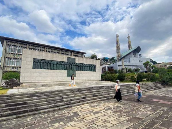 西坂公園。写真左手が「日本二十六聖人記念碑」、彫像は彫刻家･舟越保武が手掛けた。その背後に「日本二十六聖人記念館」がある。右奥、特徴的な２つの塔を持つ聖堂が「聖フィリッポ教会」。設計の今井兼次は早稲田大学教授で、日本にアントニオ･ガウディを紹介した人物としても知られる