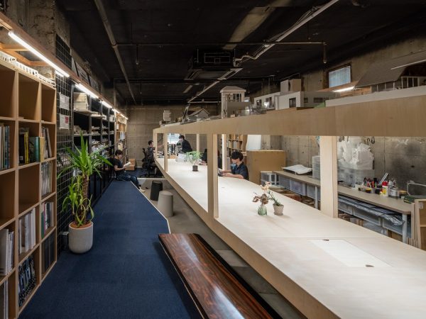 事務所の真ん中に、巨大なボックスが逆梁に乗っかっているという、不思議な空間。一見長方形のように見えるこのボックスは、奥に行くほど机の奥行が広くなる。打合せをする手前は対面で話すのに適度な距離感、奥は作業席として広さを確保。また緩やかにカーブを描き、ところどころに柱が入ることで、それぞれのスペースが部屋のように独立。さらに天井があることで部屋感も出たそうだ。ちなみに手前のベンチはスナックのカウンターを再利用したものだ
（Photo:ToLoLo studio）