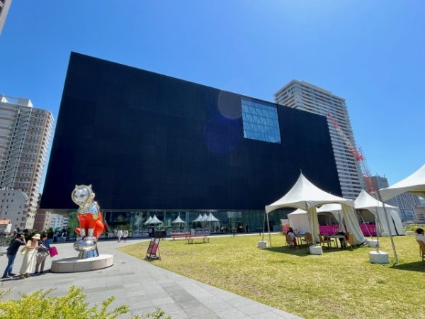 北側外観。道路面から1階分上がったところに芝生広場があり、そこからガラス張りの2階ホールにアクセスする。撮影時は「モディリアーニ ─愛と創作に捧げた35年─」（2022年4月9日〜7月18日）開催中で、これに合わせて広場でオープンカフェを営業していた。手前左のオブジェは大阪出身のアーティスト、ヤノベケンジが大阪中之島美術館のために制作した《SHIP’S CAT(Muse)》。この前で来館者が入れ替わり立ち替わり記念撮影していた