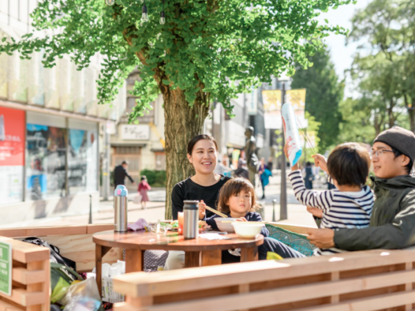 テーブルや木の周りに備え付けたベンチなどで思い思いにくつろぐ人々。足を止めて会話を楽しむ

