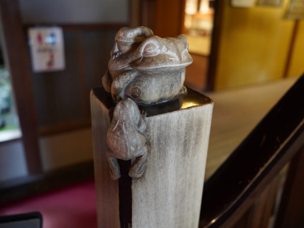 階段の親柱をかわいく彩る「二見かえる」は、彫刻家・板倉白龍の作品。昭和11年と刻銘されていることから、2度目の増改築の最後に設置されたことが分かる。別で作ったものを接着したのではなく、楠の親柱そのものに彫られている