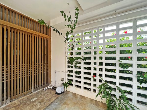 ▲“閉じつつ開く”の工夫は随所に見られる。南西側に面した駐車スペースの壁面は風通しの良い花ブロックに。その周りに植えられたハイビスカスが日よけになっている。「有孔コンクリートブロック（花ブロック）は、日中の日射しを遮断するほか、目隠し効果もあるため、防犯対策をしながら開放部を実現しています。また、風が孔を通り抜けるので、台風の時はコンクリートで強度を保ちつつ強風のバッファー（緩衝）としても重要な役割を果たします」