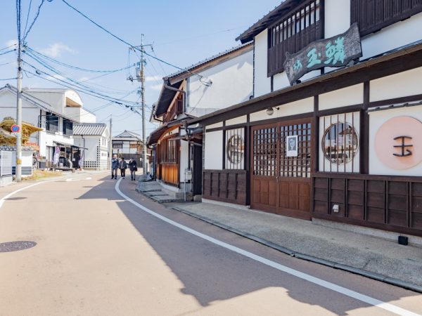港町商店街の様子。手前は茶舗「樋口芳泉園茶舗」。中央の茶色いモダンな建築は銭湯「みなと湯」だったが、現在は廃業。奧は玉島の鎮守・羽黒神社の東参道口