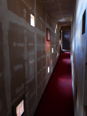 床が斜めの廊下。よく見るとドアの枠も斜め。壁に赤い光が映っているのは、廊下の右の部屋に差し込んだ光がカーペットに反射したもの。狭い廊下は雁木にも見える