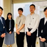 東京都庁の会議室の窓際に5名が一列に立って並んでいる。左から2人目は女性。