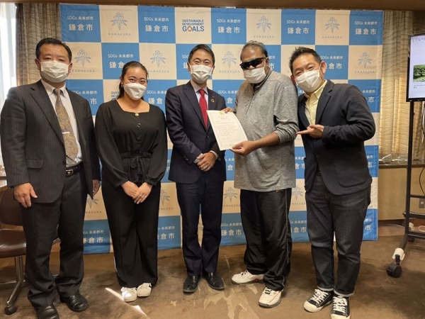 鎌倉市の地域通貨を活用して地域交流に貢献したとして、鎌倉市長から表彰状を授与されたときの様子。鎌倉市長を囲んで、アルペなんみんセンターのスタッフと、表彰状を手にした難民の方がにこやかに並んでいる