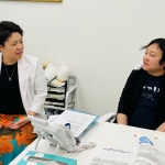 精神科の診療室にて、椅子に並んで座り、顔を見合わせて話す様子のる2名の女性。