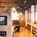 大きな窓枠や壁に造りつけられた棚が木造で、板張りの広い室内。電球色の照明が各所に室内を照らす。入口と思しき木製パーテーションの一か所に、黒い正方形の板に白い塗料でソーシャルスクウェアーのロゴがあしらわれている。