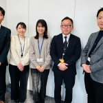 白い室内の壁を背景に、5名が立って並んでいる。
