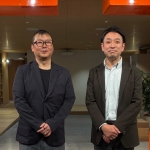 オフィスのエントランスで並んで立つ2人の男性。左の男性は眼鏡をかけている。